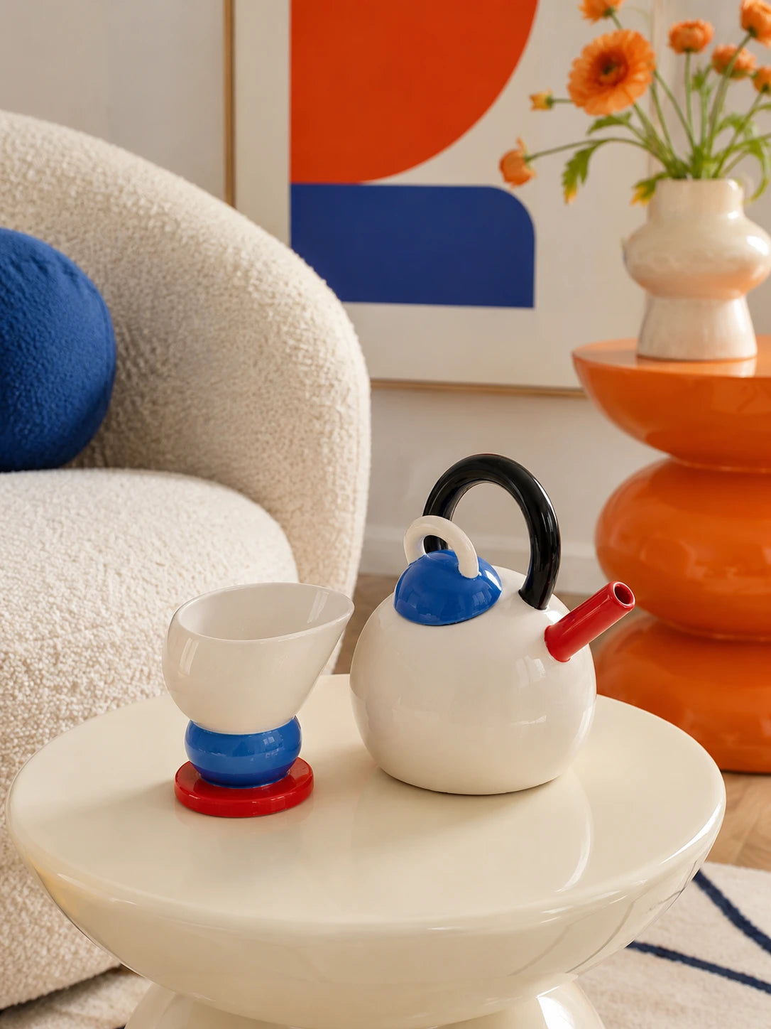 sphere ceramic teapot and cone cup set on a cream side table by a boucle armchair with an orange stool behind