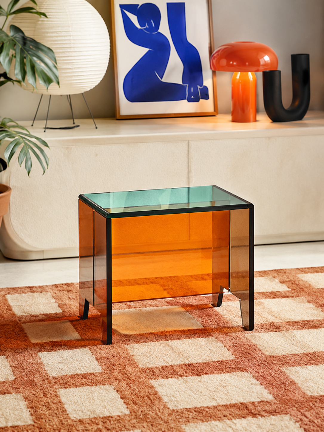 orange acrylic mini side table on a checkerboard rug in front of a cream sideboard with a paper lantern and orange lamp