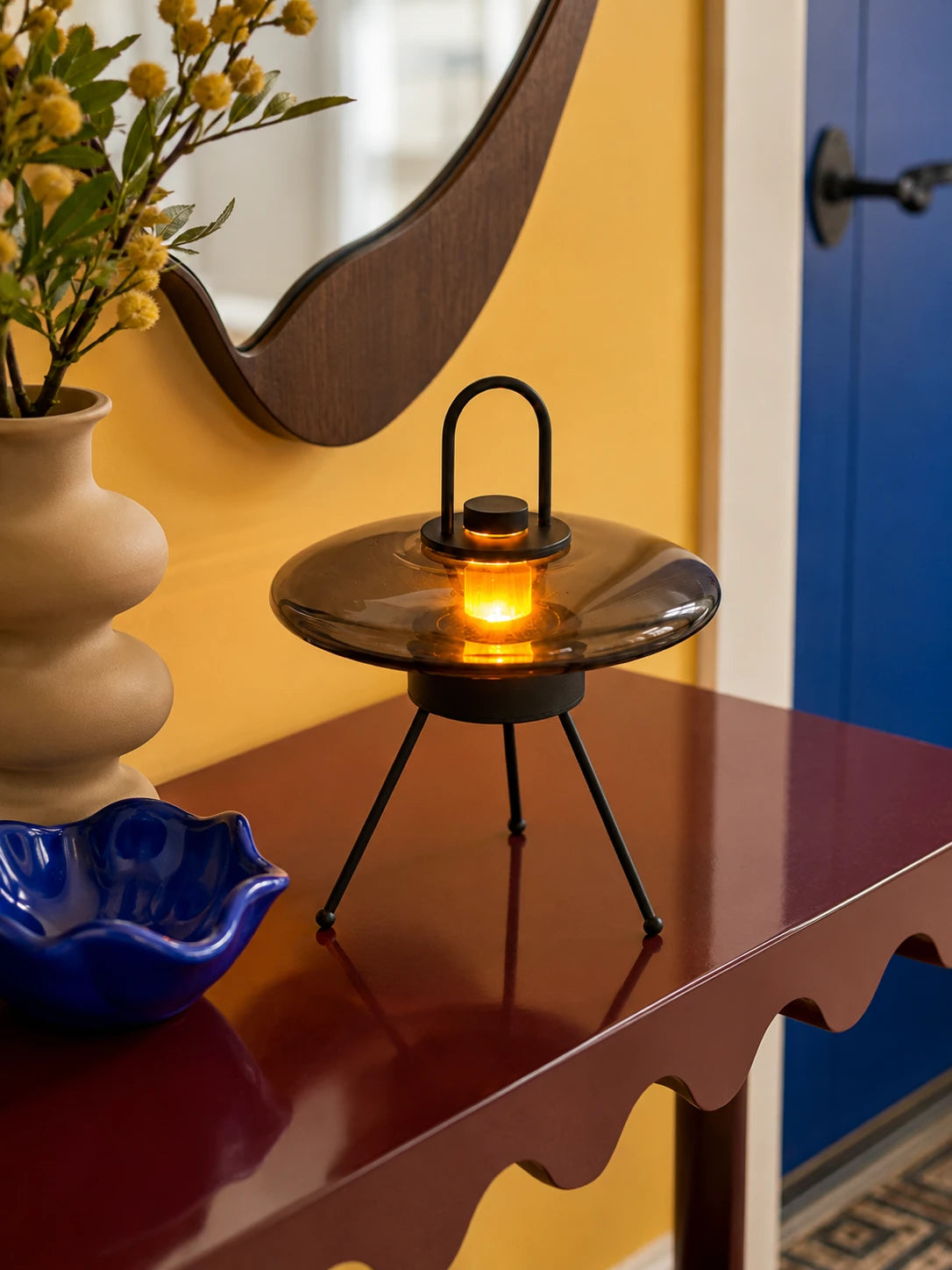 Smoked glass saucer lantern lamp lit warm on a burgundy console under a wavy wood mirror