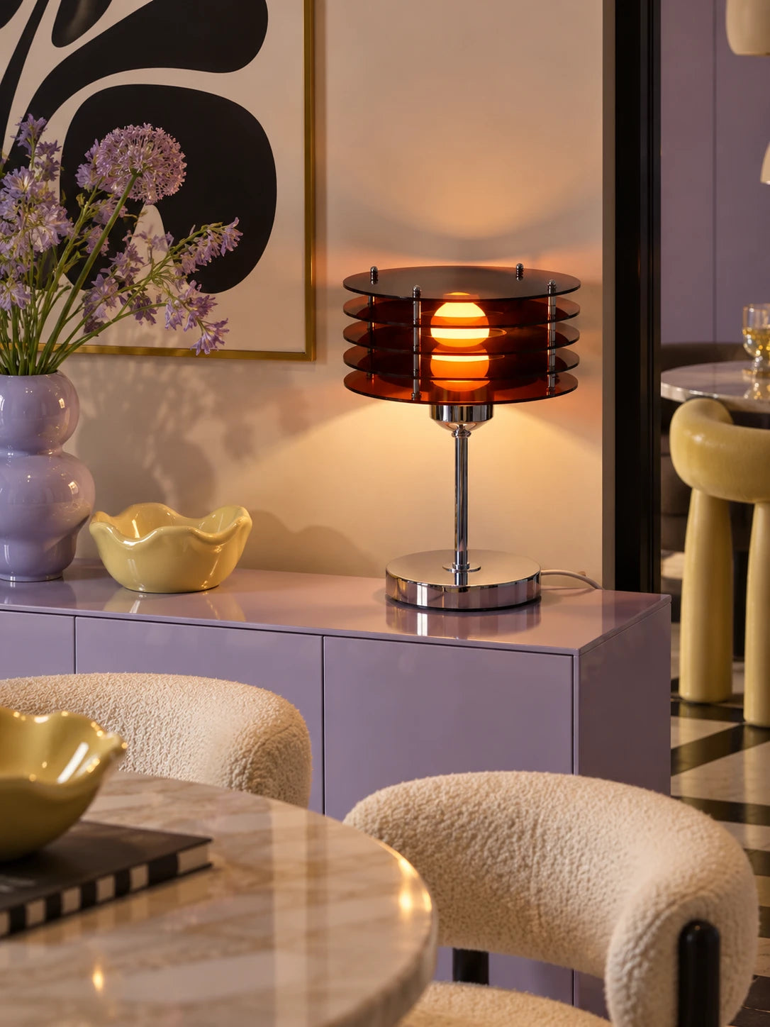 Smoked acrylic disc lamp lit on a lilac sideboard with a yellow wavy bowl and purple allium flowers