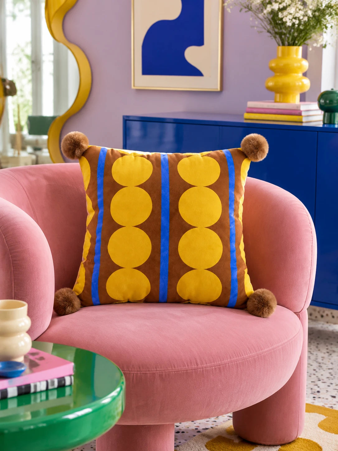 Brown and yellow dot stripe pillow with pompom corners on a pink velvet armchair, lilac wall behind