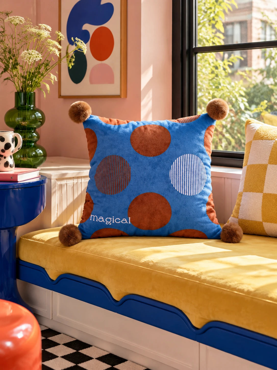 Cobalt polka dot pompom pillow on a mustard window seat with a yellow checkered pillow behind