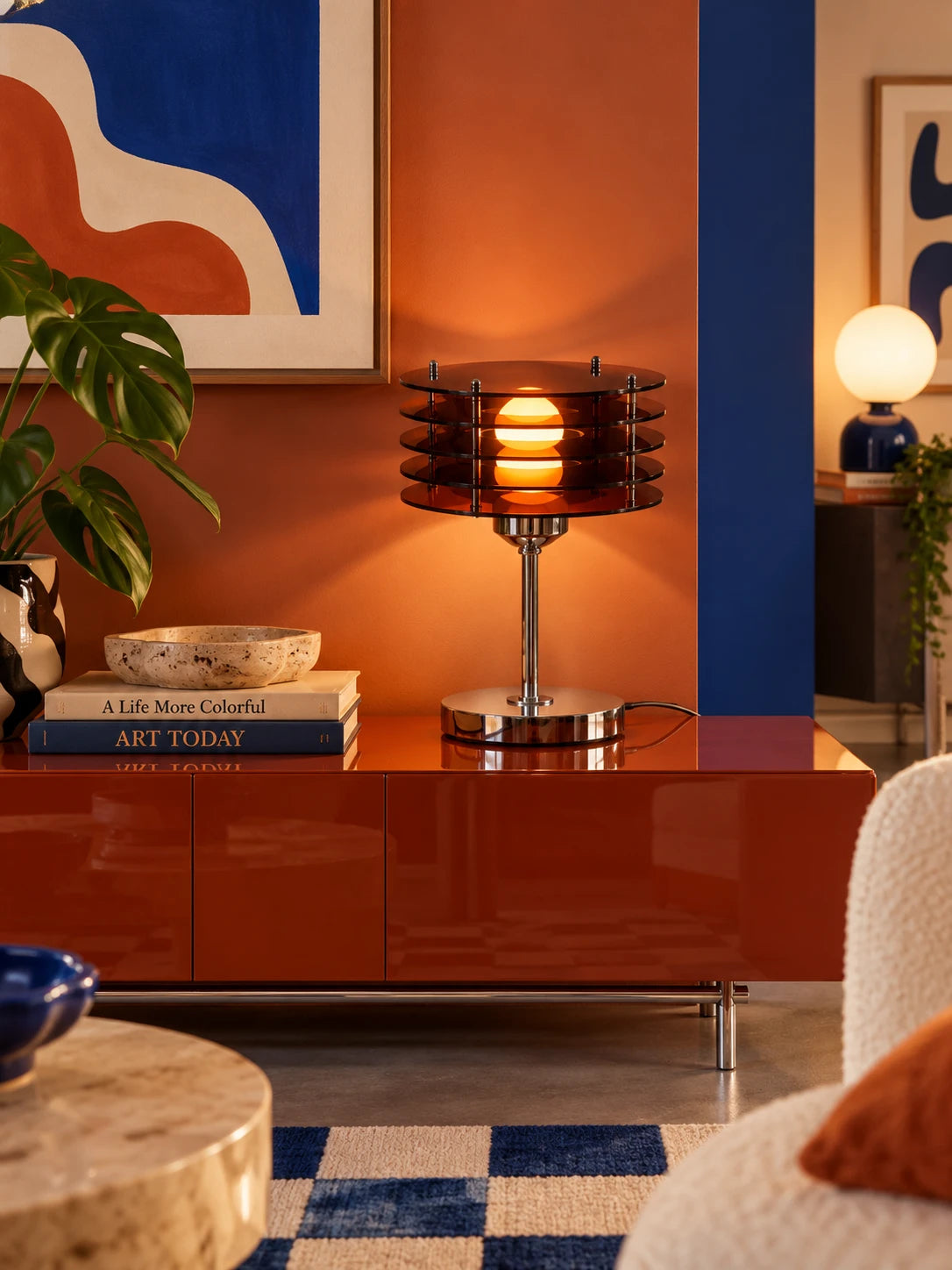 Acrylic disc table lamp on a glossy rust media console with art books, a monstera and a checkered rug