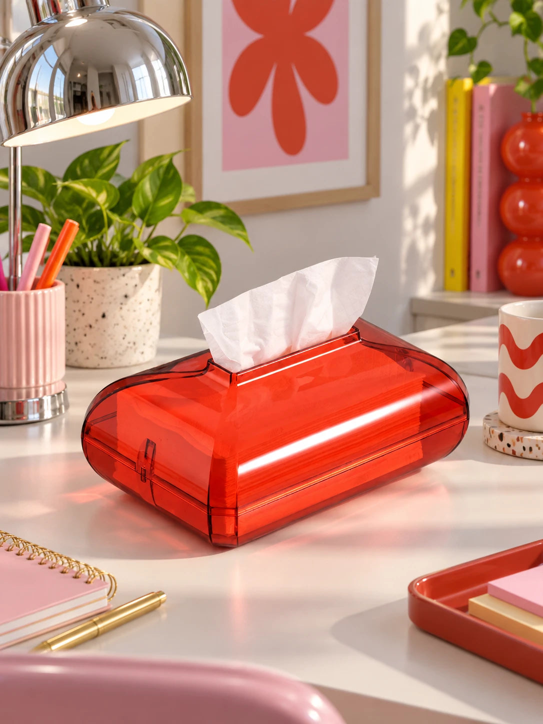 Red acrylic tissue box on a desk with a chrome lamp and notebooks