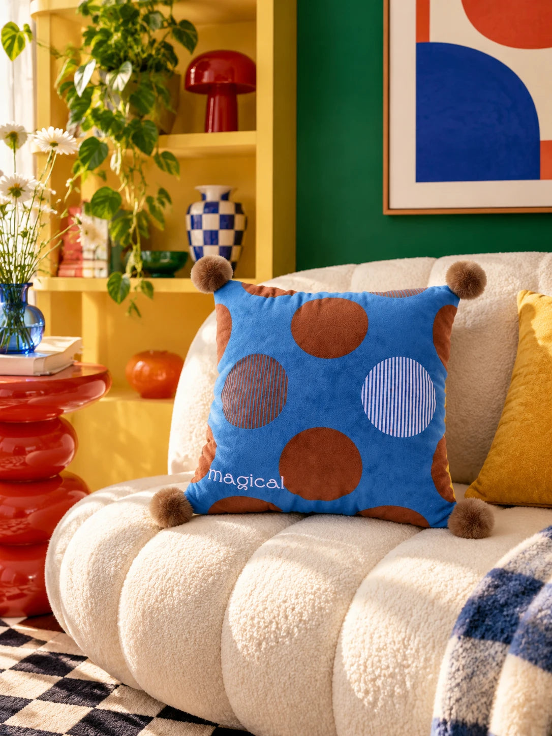 Cobalt and brown polka dot pompom pillow on a cream channel sofa by yellow shelves and a green wall