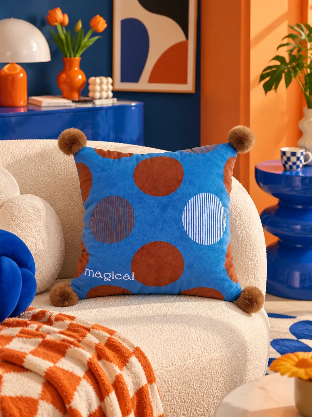 Cobalt polka dot pillow with brown pompoms on a cream boucle sofa beside an orange checked throw