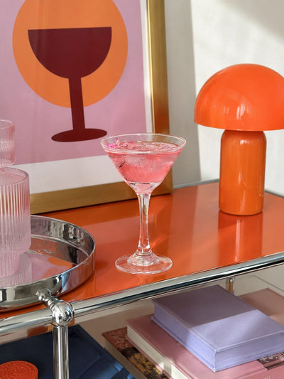 pink cocktail in a clear martini glass on an orange bar cart beside an orange mushroom lamp