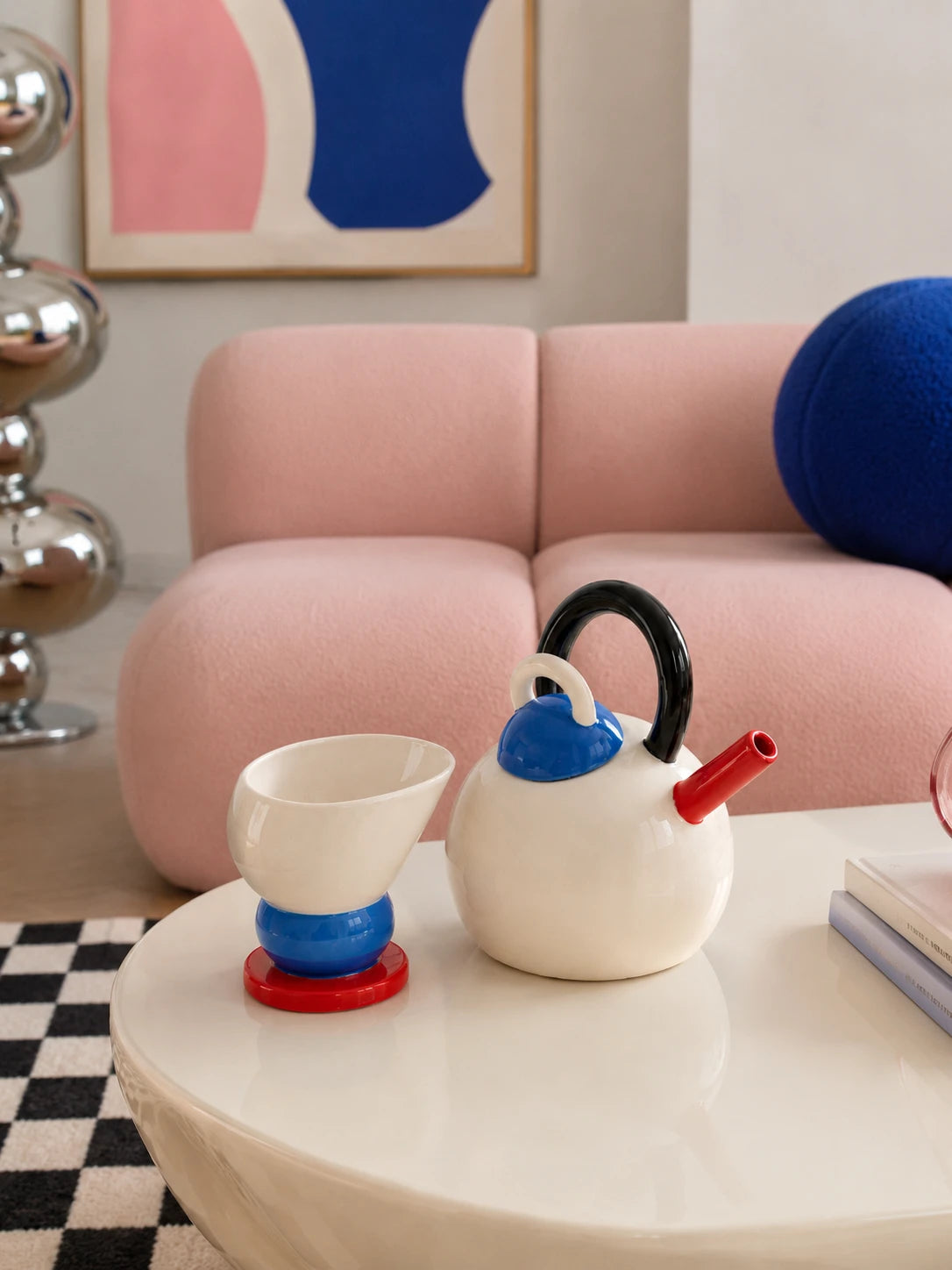 cream ball teapot and stacked cone cup on a white coffee table in front of a pink sofa and a checkerboard rug
