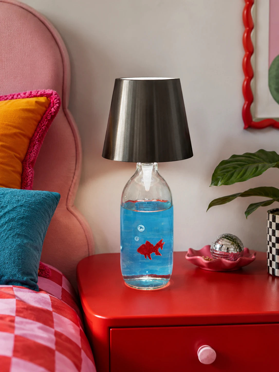 goldfish bottle lamp glowing softly on a red nightstand beside a pink upholstered headboard