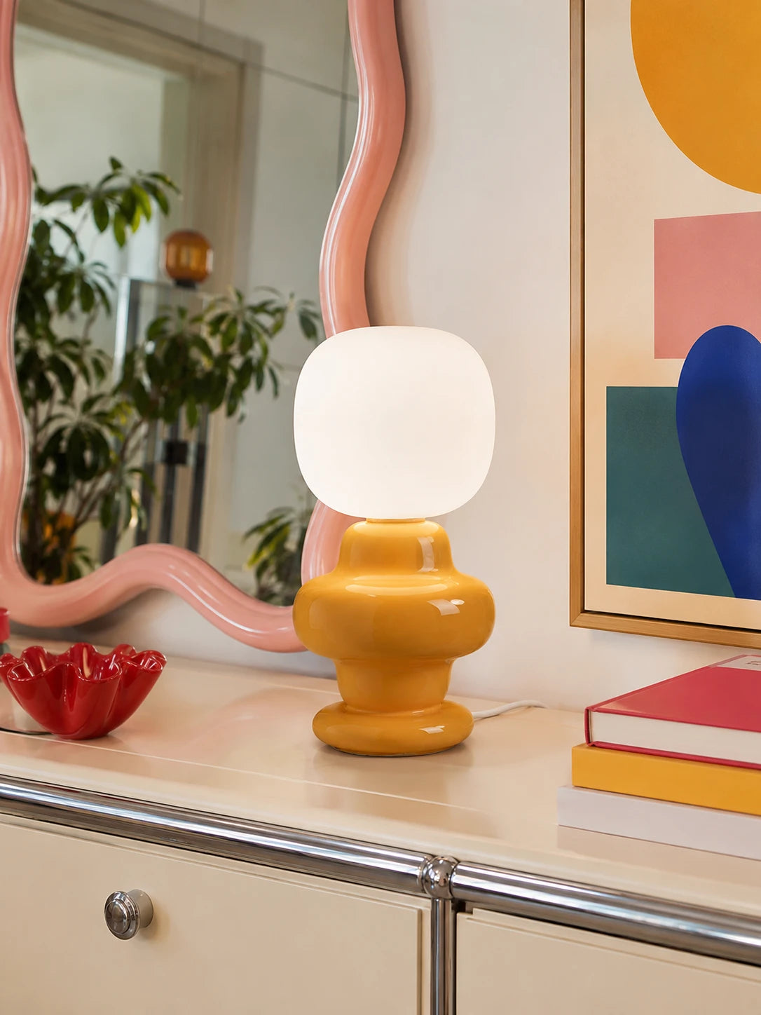 yellow ceramic Memphis table lamp glowing on a cream console beside a wavy pink mirror