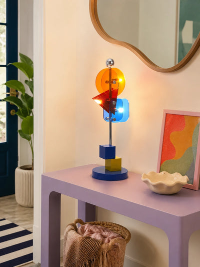 Memphis geometric lamp lit on a lilac console table beneath a wavy wood-framed mirror