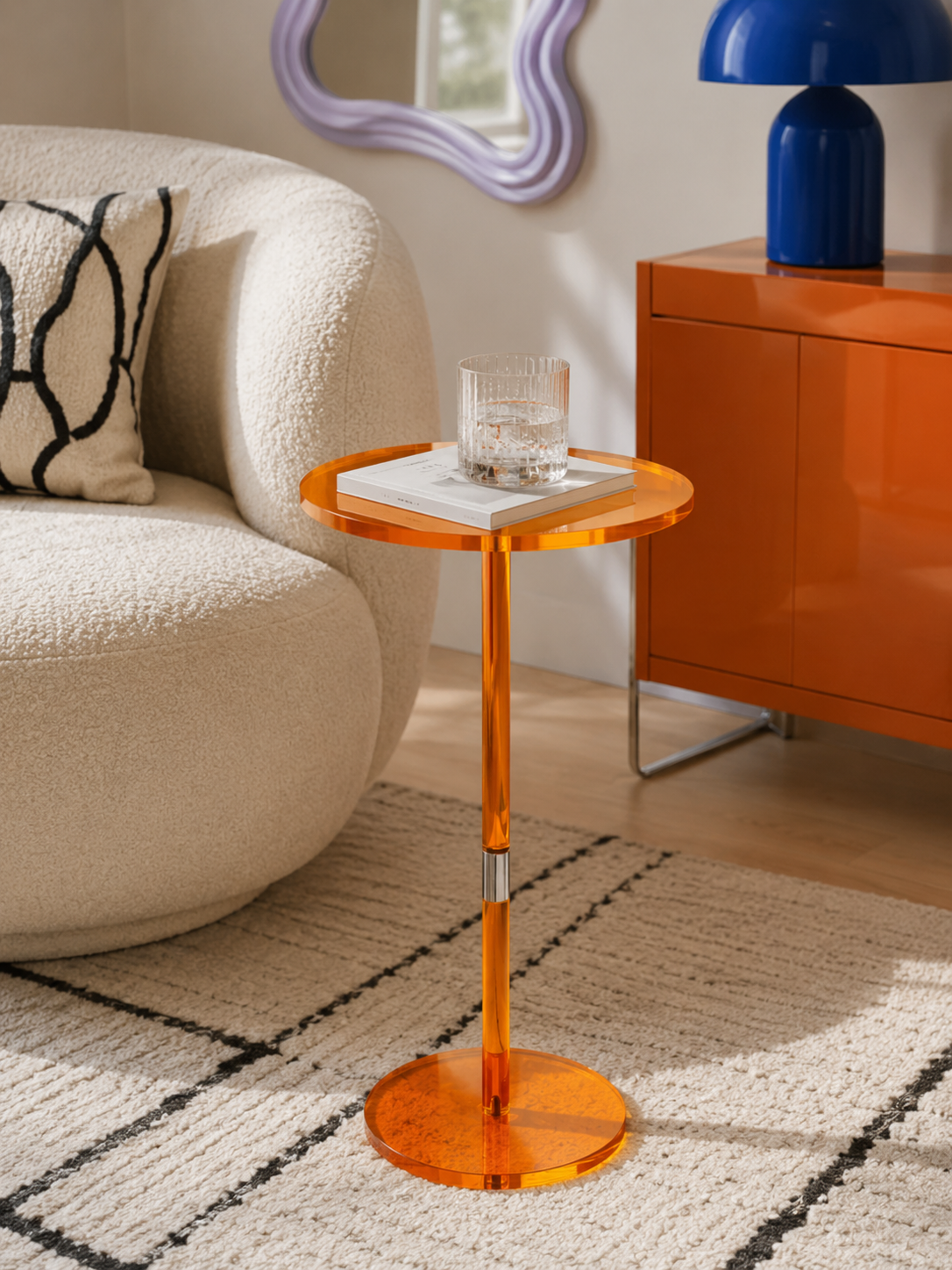 orange acrylic pedestal side table holding a glass and a book beside a cream boucle sofa and an orange cabinet