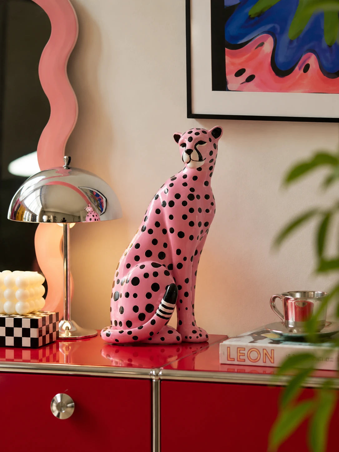 pink polka-dot leopard statue on a red cabinet beside a chrome mushroom lamp and a wavy pink mirror
