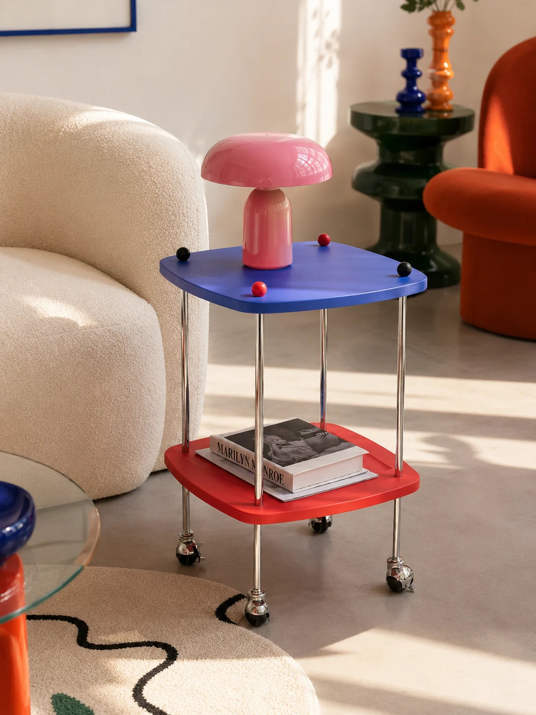 blue and red two-tier rolling side table holding a pink mushroom lamp beside a cream boucle sofa