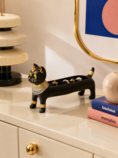 black and gold Egyptian cat jewelry tray on a cream dresser next to stacked books and a gold-framed mirror