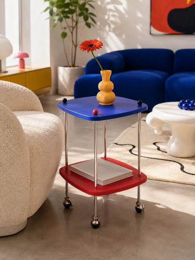 cobalt and red two-tier side table on casters with a yellow vase and orange flower in front of a blue sofa
