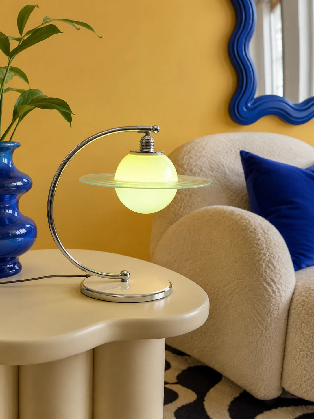 Glowing green planet lamp with a glass orbit ring on a cream side table beside a blue vase and boucle chair