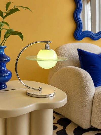 Glowing green planet lamp with a glass orbit ring on a cream side table beside a blue vase and boucle chair