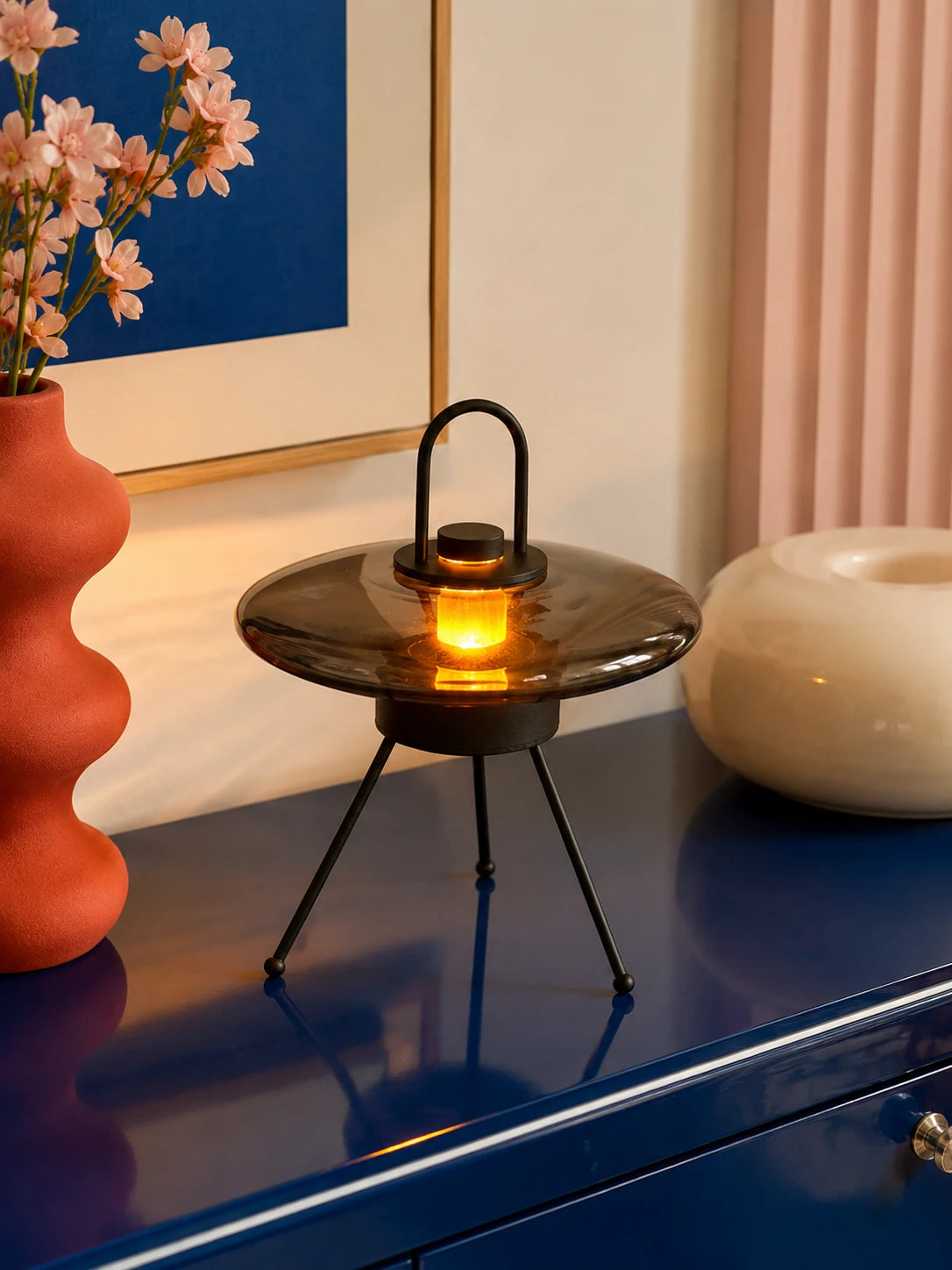 UFO tripod lamp glowing warm amber through a smoked glass saucer on a blue sideboard beside a red vase
