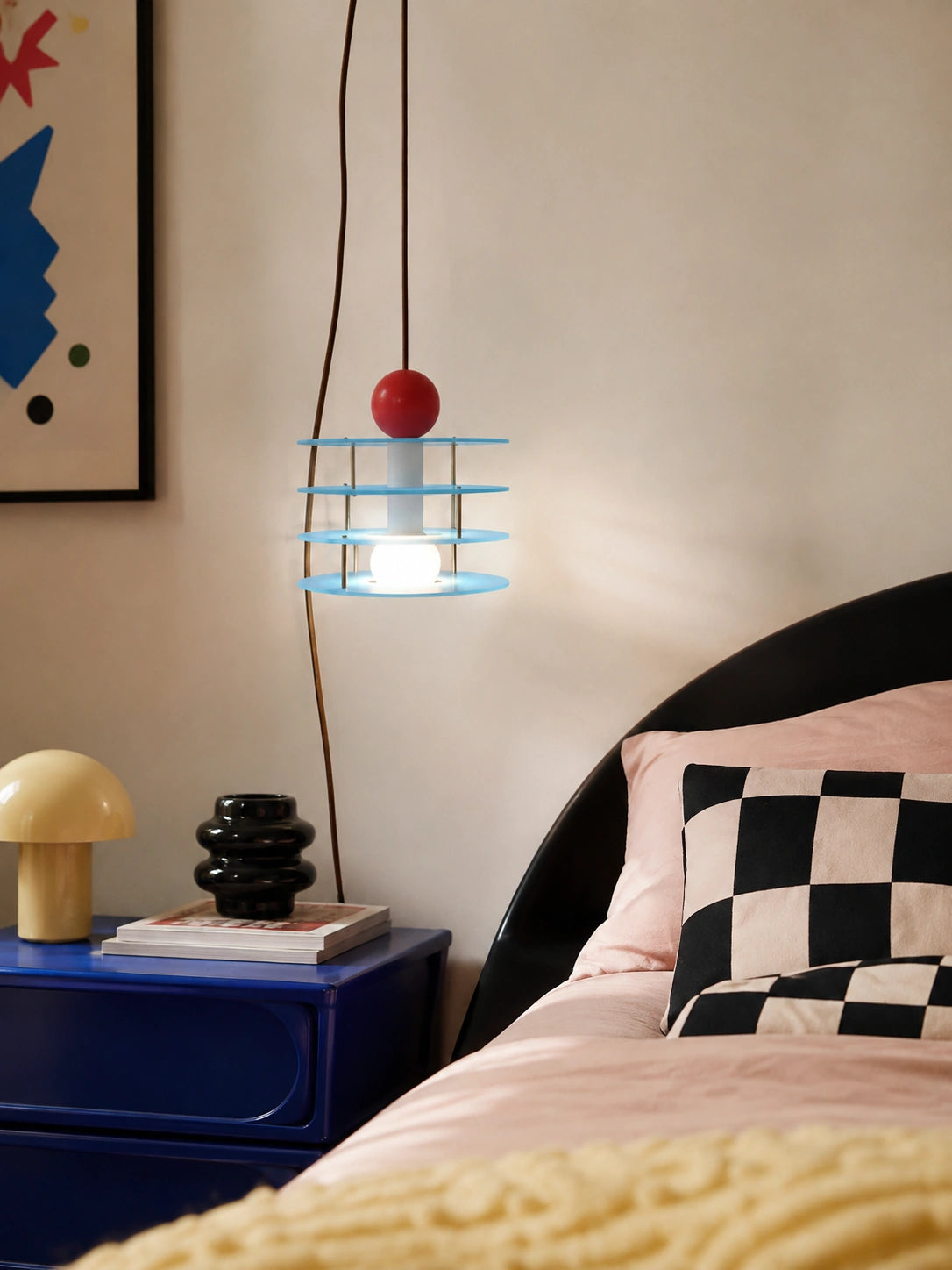 blue macaron pendant light glowing beside a bed with pink bedding, checkered pillow, and cobalt nightstand