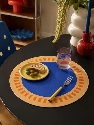 blue and tan sun-dial placemat on a black round table with a yellow plate of olives, a fork and a red vase