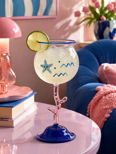 coral stem cocktail glass holding an iced drink with a lime wheel on a pink side table beside a blue armchair