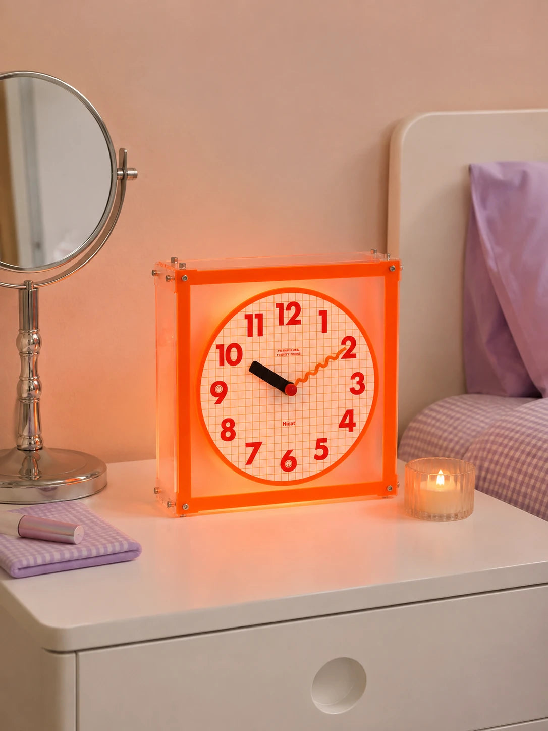 orange grid dial clock on a white nightstand beside a lit candle and a silver vanity mirror