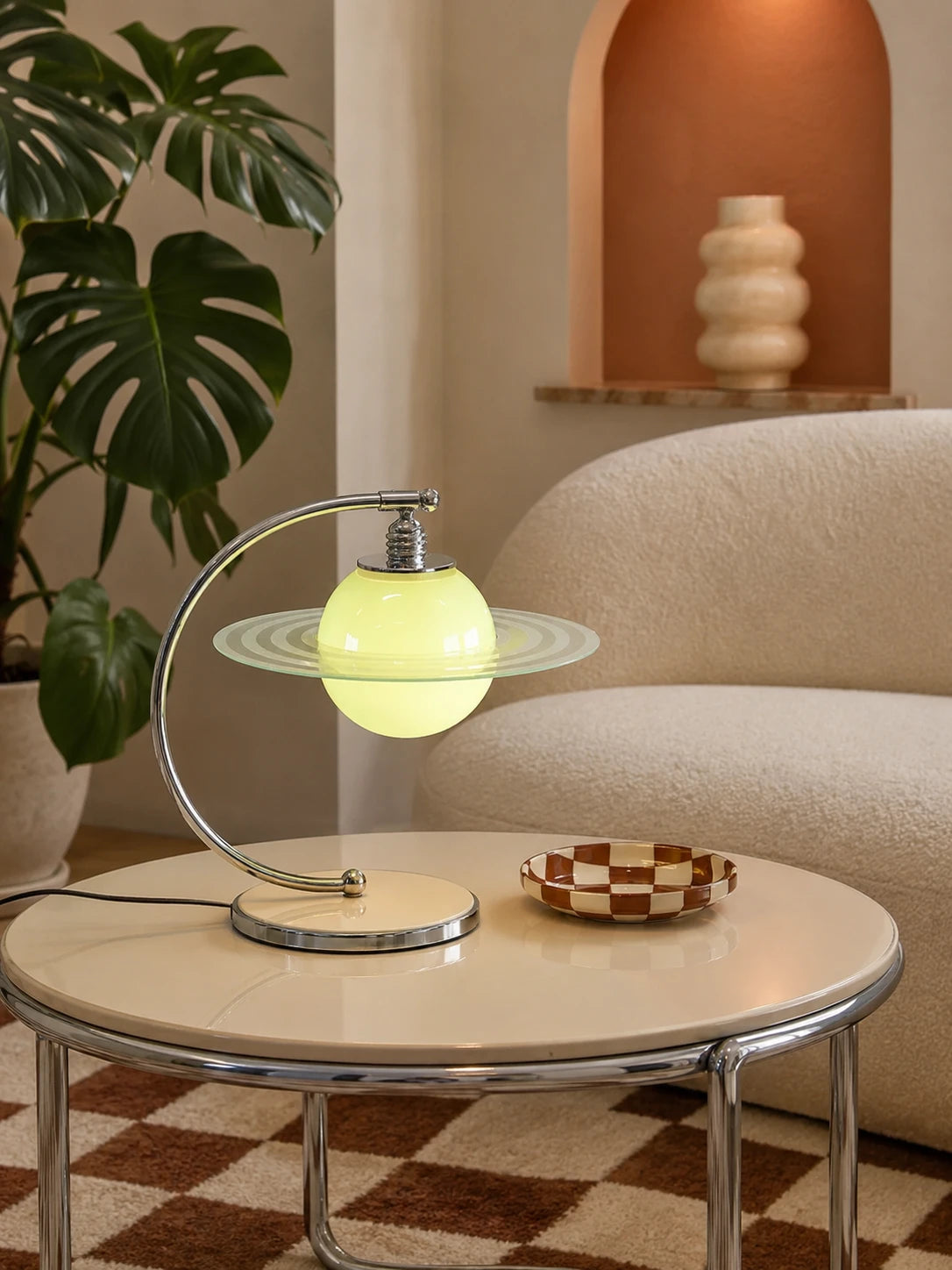 Saturn planet table lamp glowing on a cream coffee table with a checkerboard dish and a monstera plant