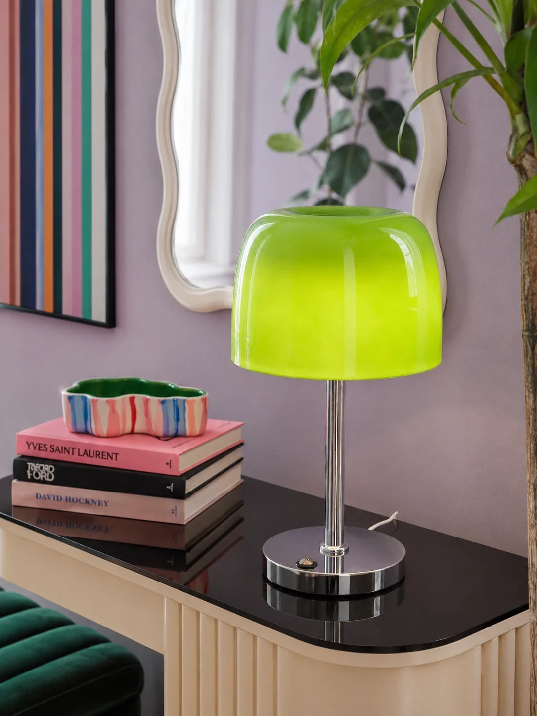 Green glass mushroom lamp on a black console with stacked art books, a wavy mirror and a potted plant