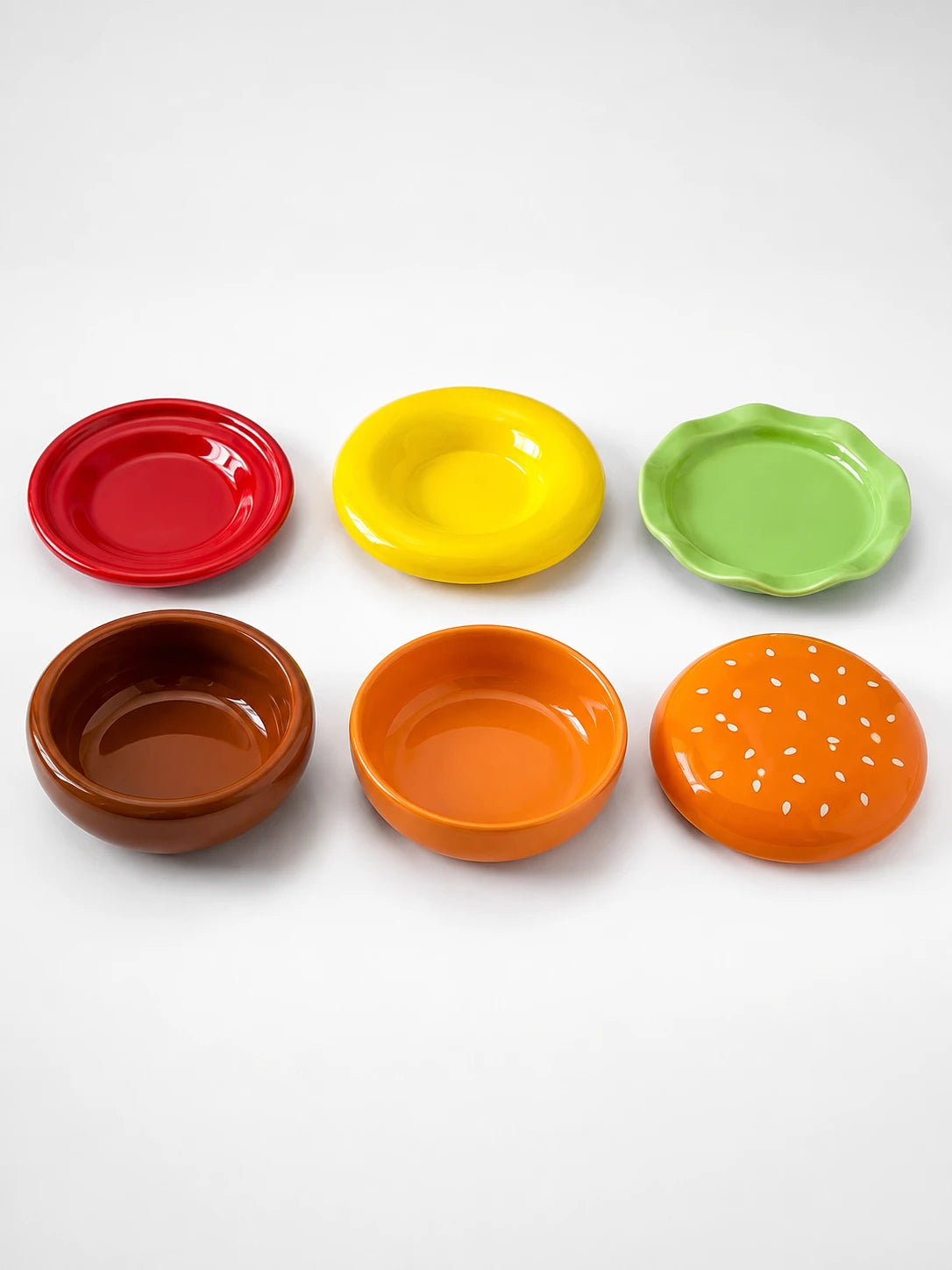 six ceramic hamburger plate set pieces laid out: red, yellow and green plates, brown and orange bowls, sesame bun lid