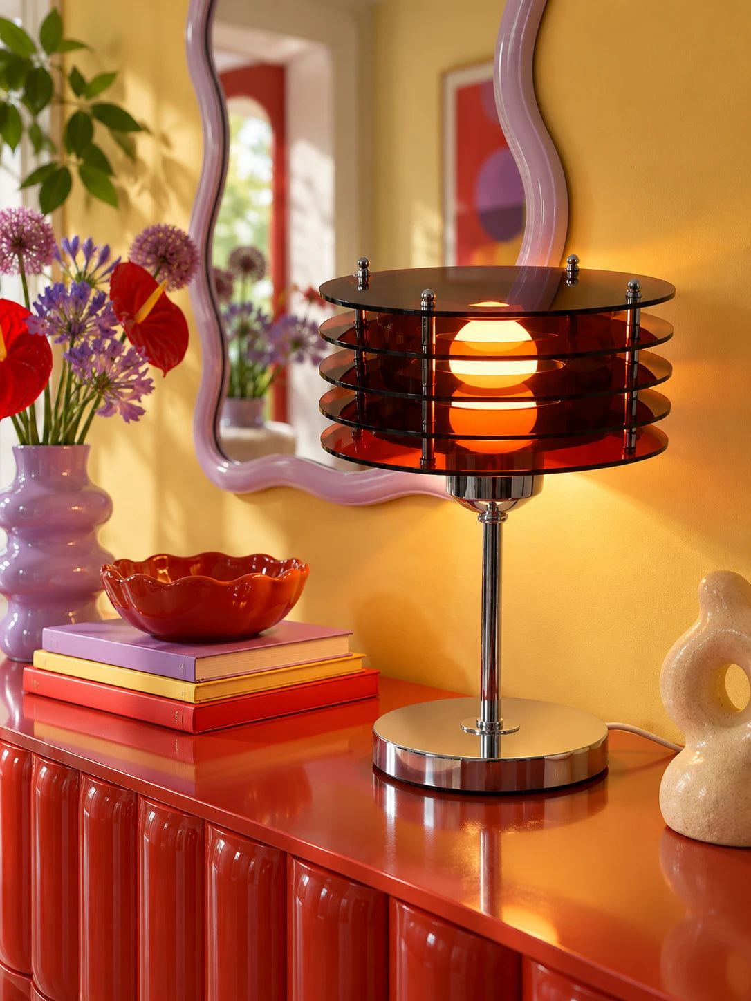 Amber disc table lamp on a red ribbed sideboard by a lilac wavy mirror and red anthurium flowers