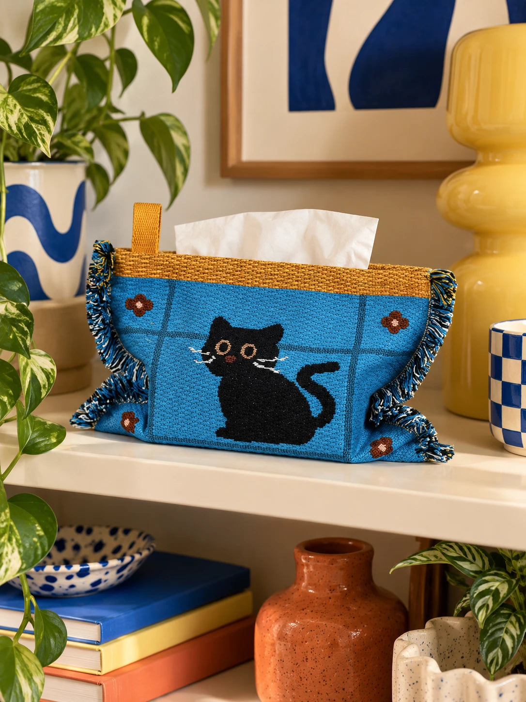 
blue cat knitted tissue pouch on a wood shelf beside books and a plant