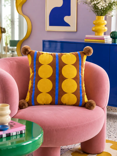 Brown and yellow dot stripe pillow with pompom corners on a pink velvet armchair, lilac wall behind