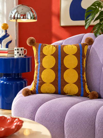 Brown and yellow dot stripe pompom pillow on a lilac channel armchair beside a cobalt side table