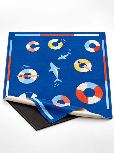 Cobalt pool party swim ring area rug with sharks and floating swimmers, top layer folded back over a black anti-slip base