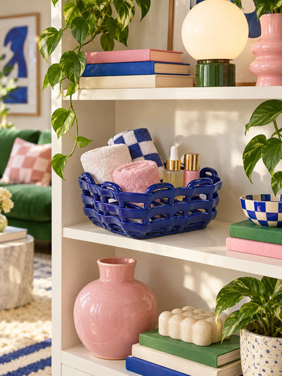 Woven blue ceramic basket with towels on a white shelf beside a pink vase