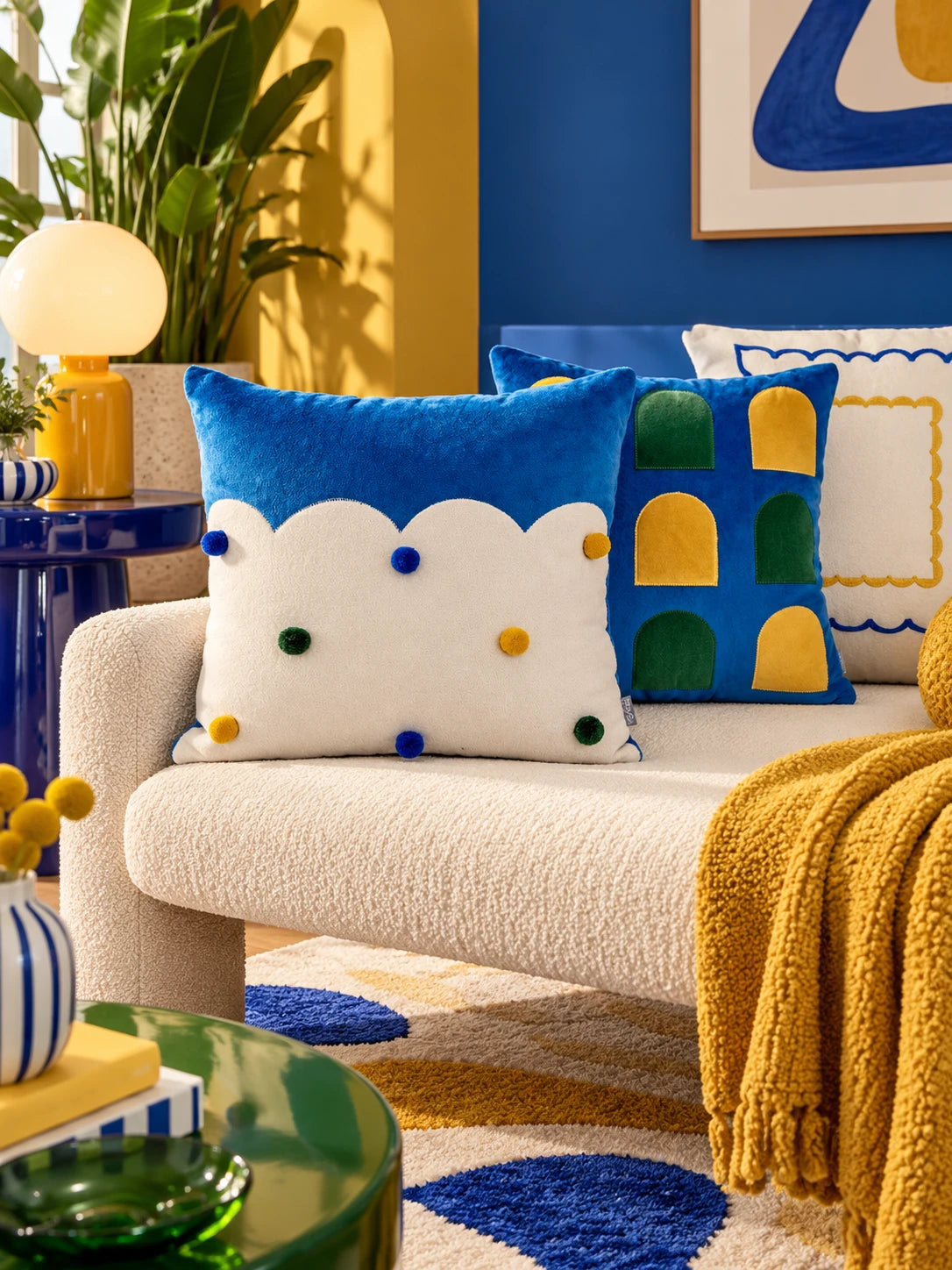 Cloud-edge pompom pillow in front of a window-arch pillow on a cream sofa with a mustard knit throw