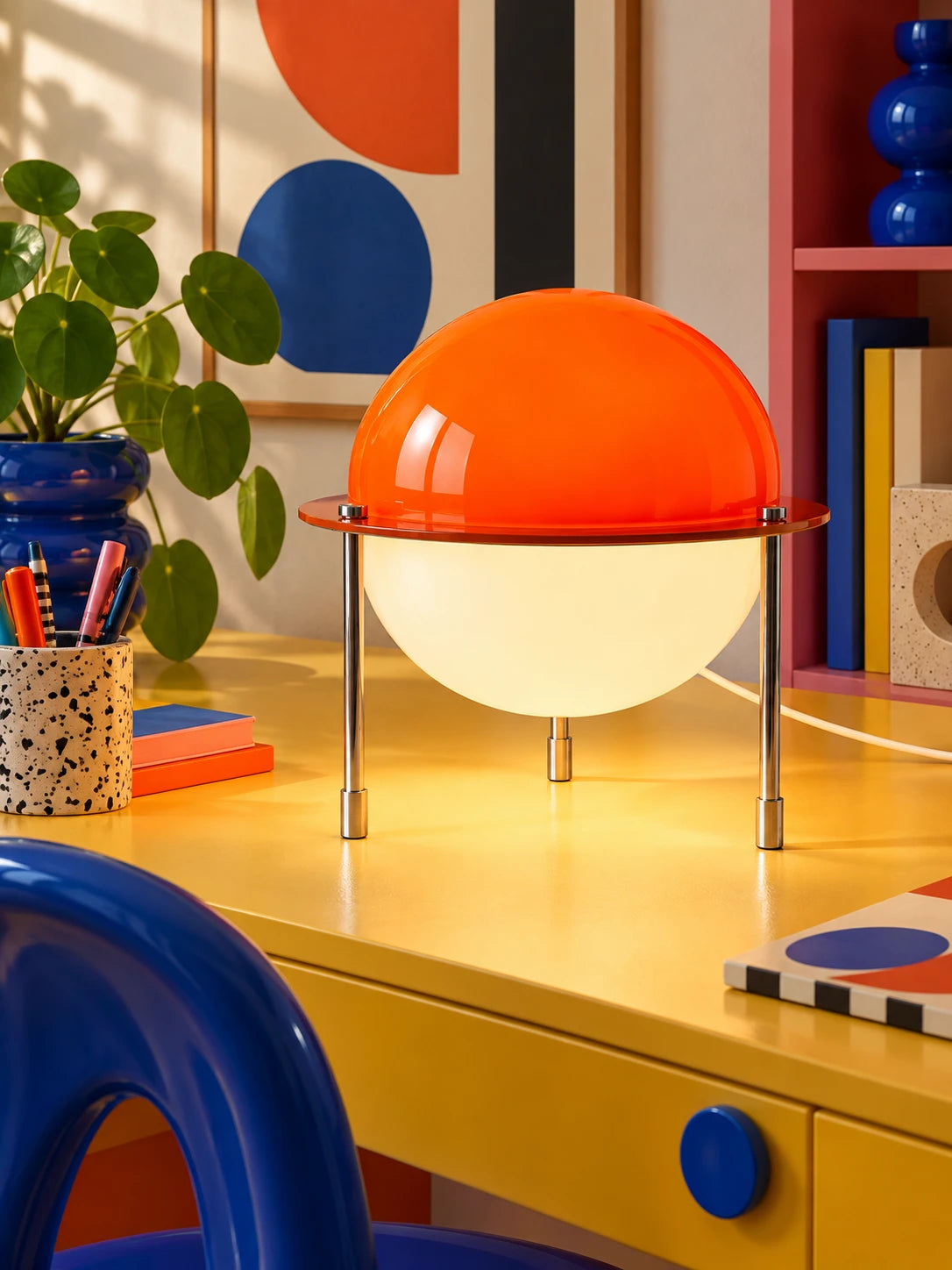 Split-sphere desk lamp glowing on a yellow desk beside a speckled pen cup and blue chair