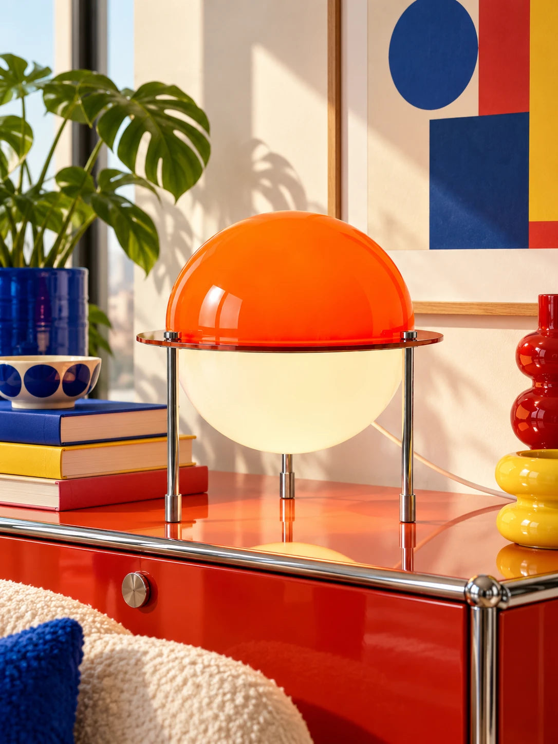 Orange split-sphere table lamp on a red sideboard with stacked books and blue plant pot
