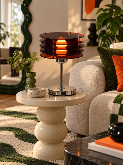 Amber acrylic disc lamp on a cream stacked side table next to a green velvet pillow and wavy rug