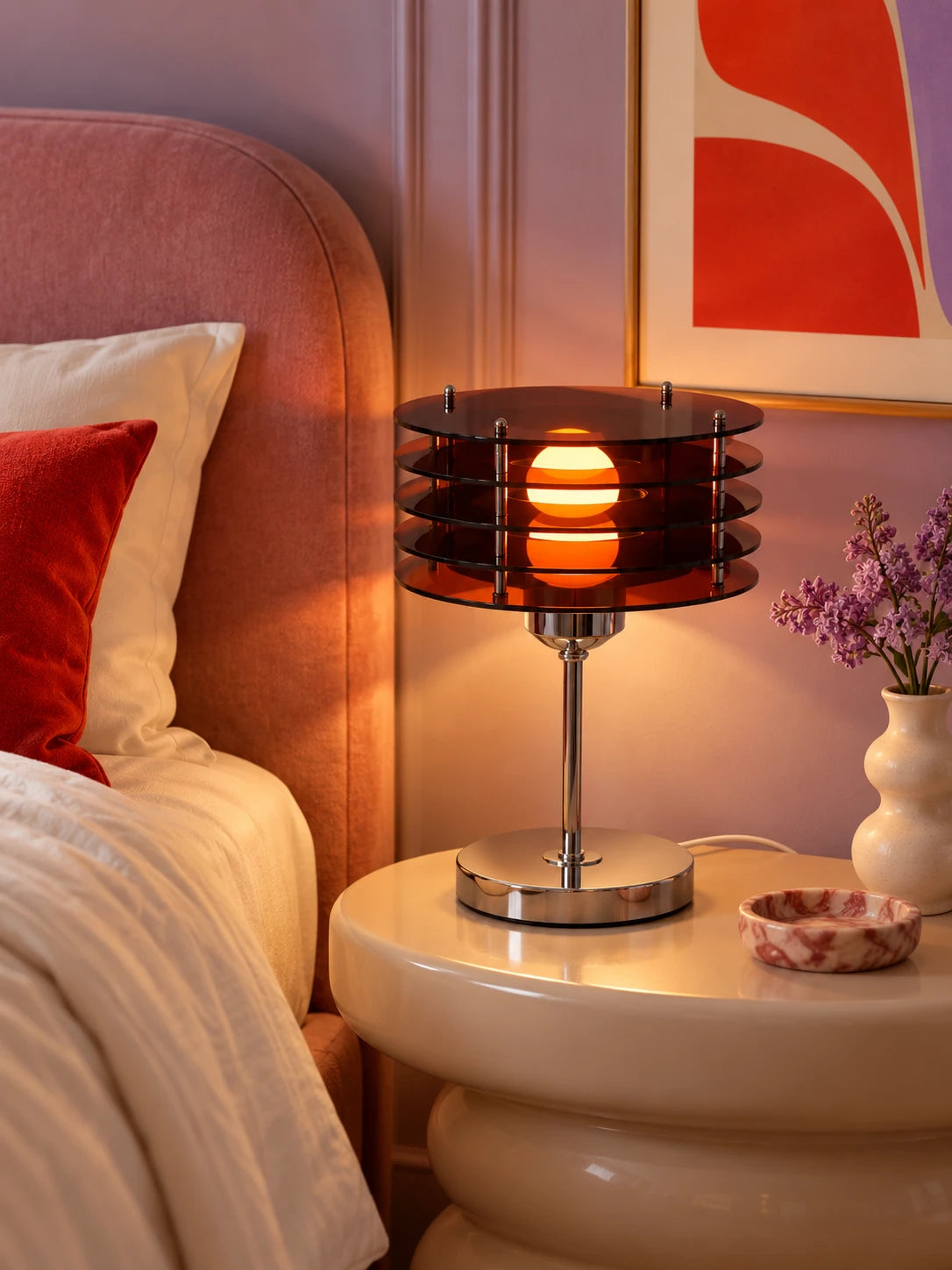 Smoked acrylic disc table lamp on a cream nightstand beside a pink upholstered bed and lilac flowers