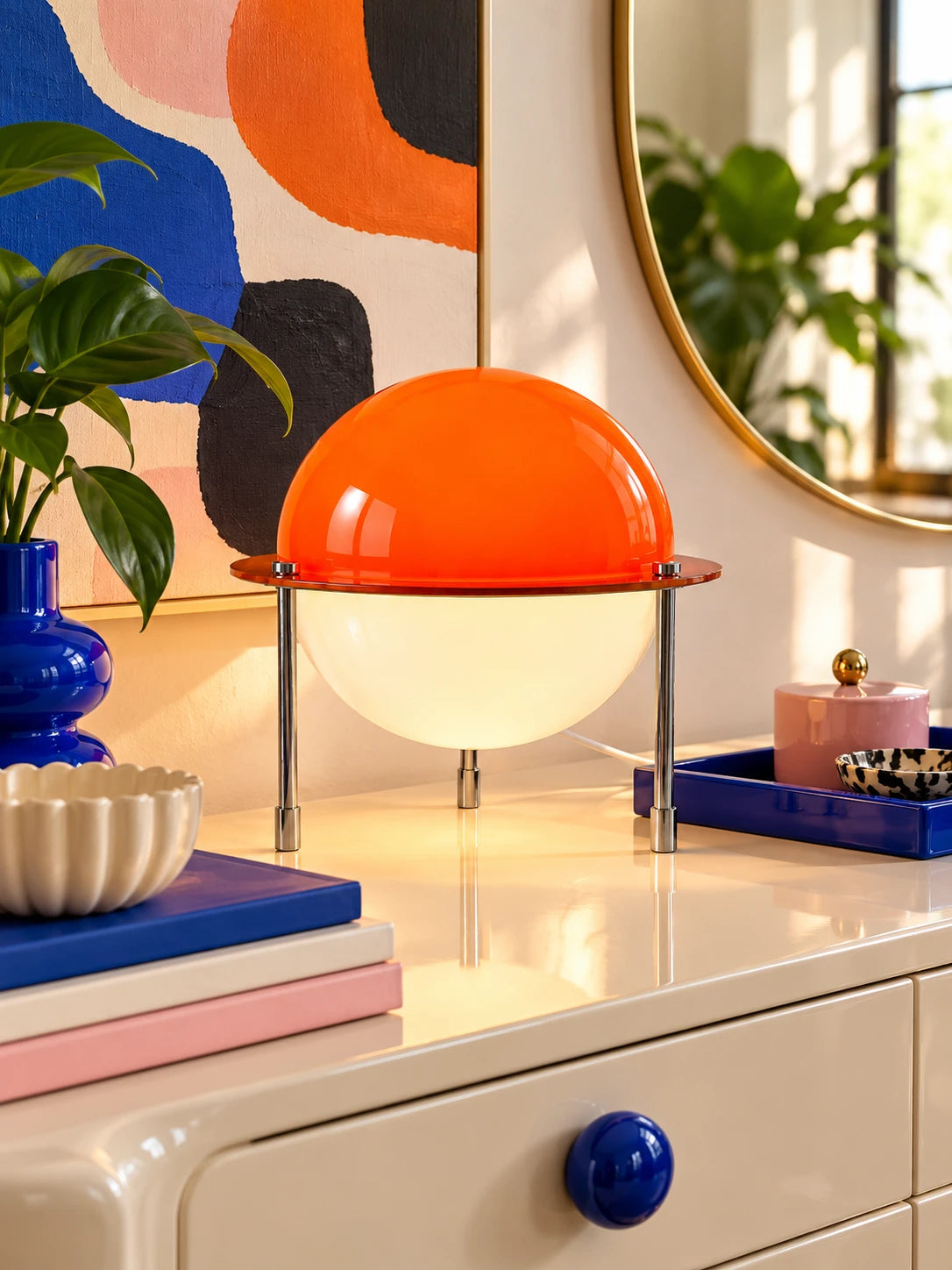 Orange dome lamp lit on a dresser beside a gold round mirror and blue trinket tray