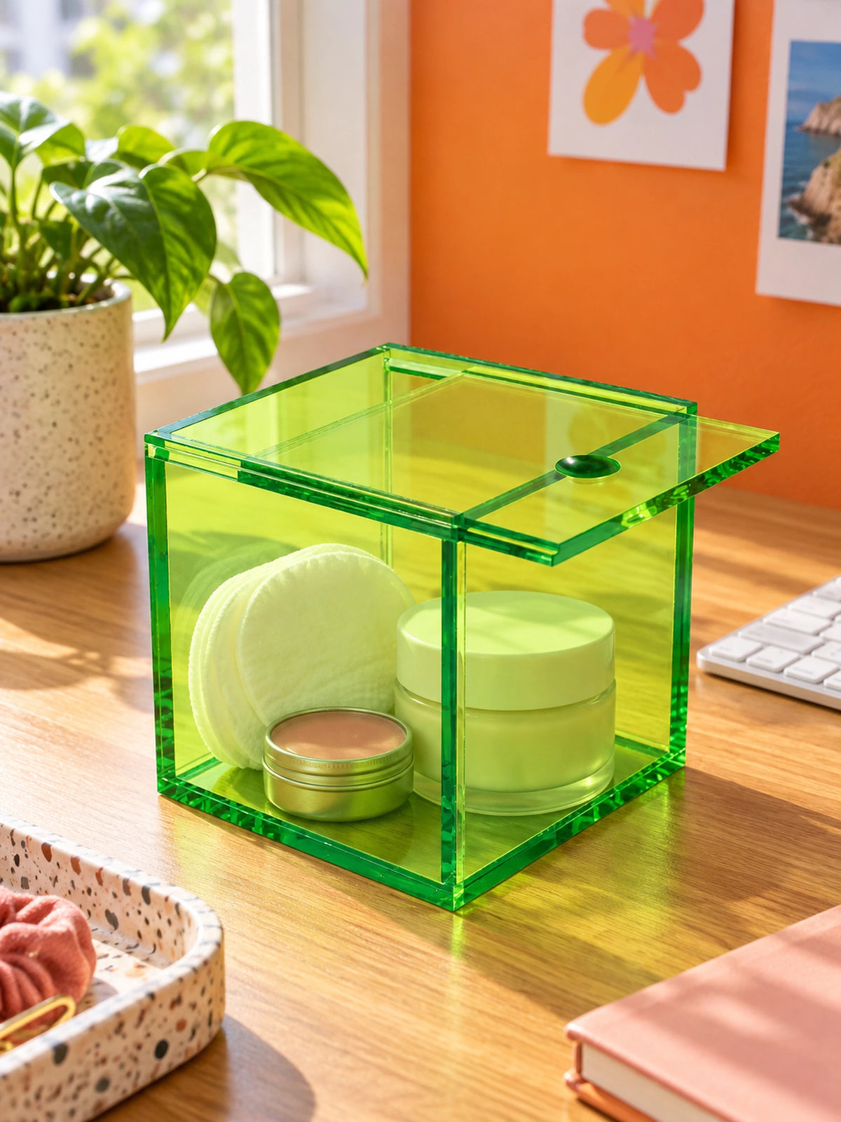 green acrylic sliding-lid box with skincare jars and cotton pads on a wood desk
