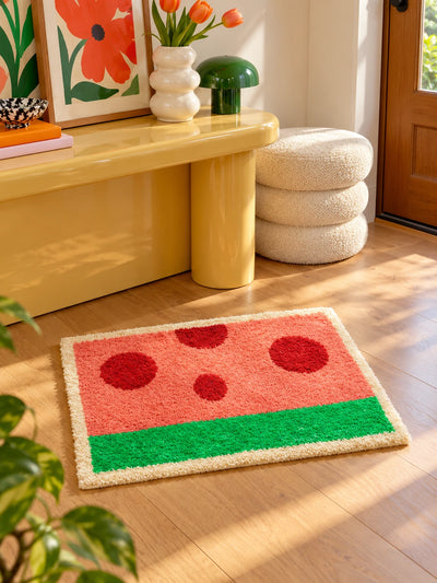 watermelon flocked bath mat on a wood floor in a sunny entryway, in front of a glossy yellow console and stacked cream boucle poufs
