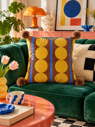 Brown and yellow dot stripe pompom pillow on a green velvet sofa with an orange mushroom lamp