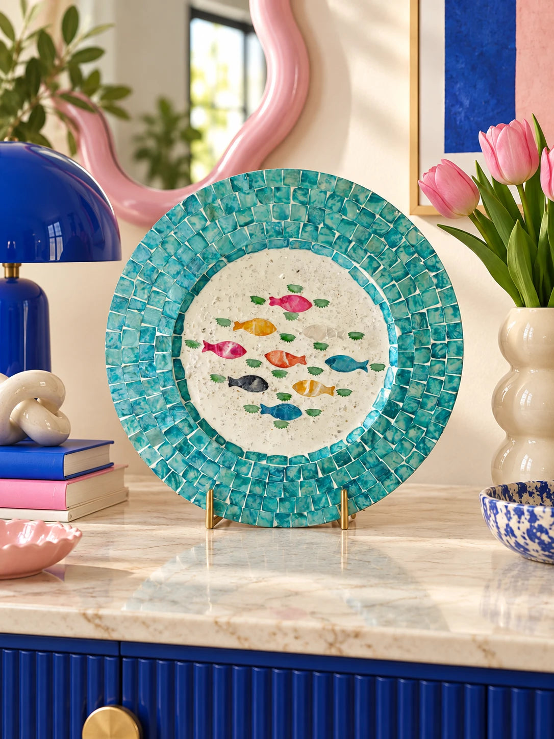 
Seashell mosaic fish tray on a marble sideboard with a pink wavy mirror and blue lamp