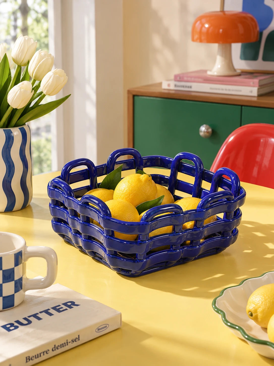 Blue woven ceramic basket of lemons on a yellow table beside a striped vase