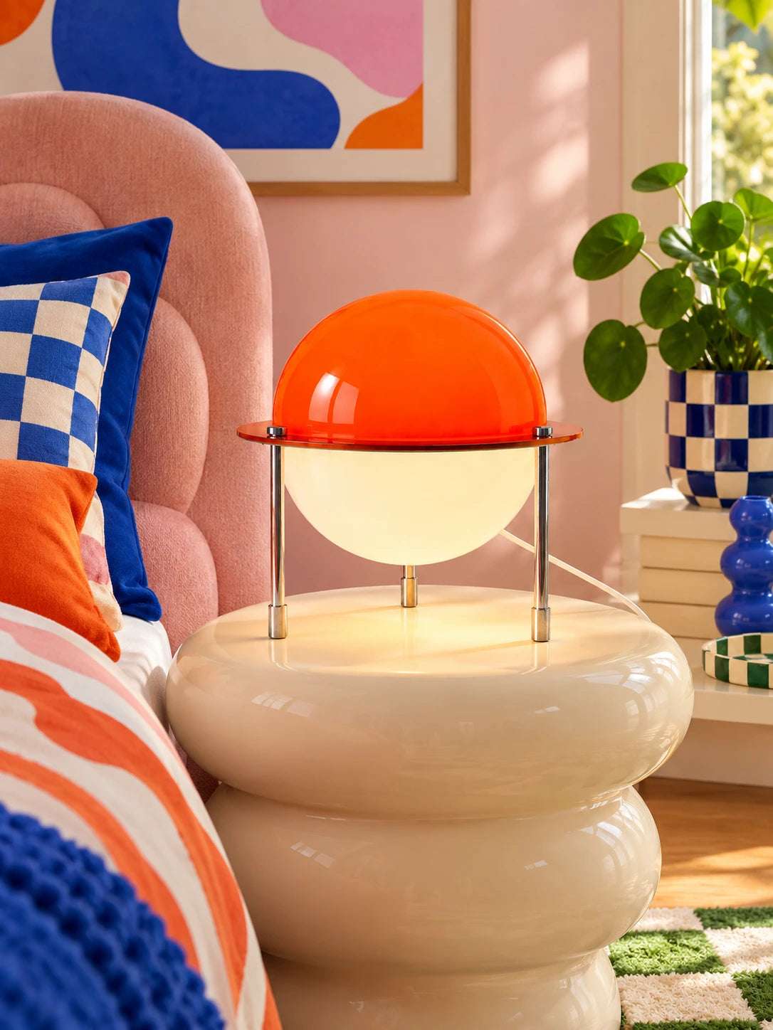 Orange dome table lamp lit on a cream bobbin side table beside a pink upholstered bed