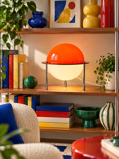 Split-sphere table lamp glowing on a wood bookshelf between colorful books and a green orb