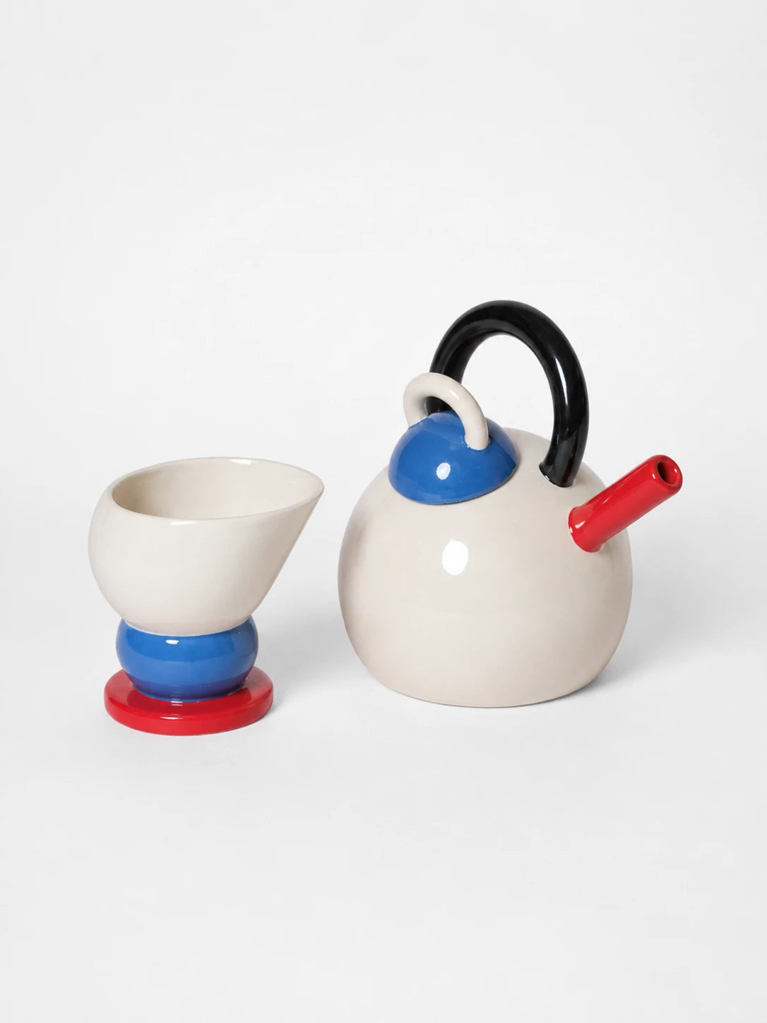 cream sphere ceramic teapot with black arch handle, blue dome lid and red spout beside a cone cup on a blue ball